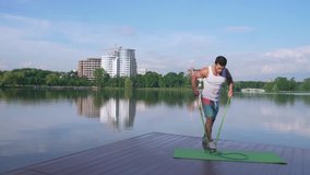 Active sportsman wearing white shirt, shorts and cap training arms with rubber expander on green yoga mat on lake pier. Fitness guy during every day workout on fresh air