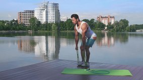 Athletic and muscular man wearing shirt, shorts and sneakers in half bent position stretching rubber band on wooden lake pier. Fitness guy doing morning exercises outdoor