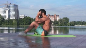 Side view of handsome fitness model in shorts doing abdominal crunches on green yoga mat near the lake during summer days. Shirtless young man enjoying workout outdoor