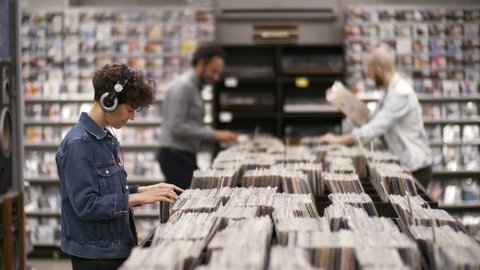Medium shot of multiethnic customers browsing large assortment of cds and vinyls in record store, chatting and showing each other discs, while curly Caucasian woman in headphones is swaying to music
