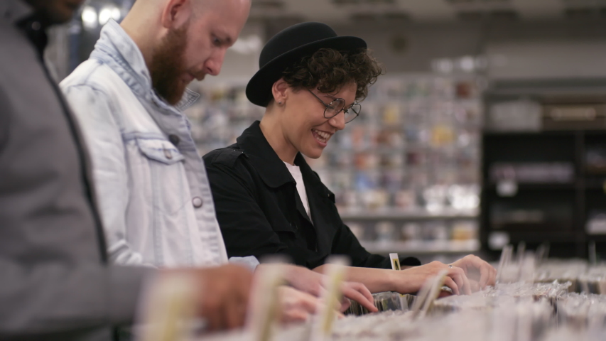 Portrait arc shot of multiethnic customers browsing through vinyls in small independent record shop, and cheerful stylish Caucasian woman finding rare disc in plain white sleeve and smiling for camera | Shutterstock HD Video #1035165431