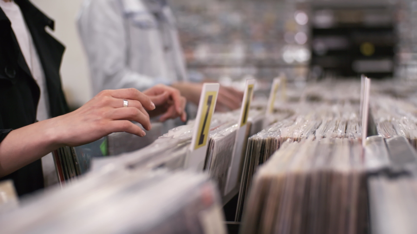 Trucking close-up hands shot of casually dressed unrecognizable female and male customers leisurely perusing cds and vinyl discs, grouped in alphabetical order, in small record shop | Shutterstock HD Video #1035167690