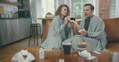 Sick Couple Sitting on Sofa in Blankets in Living Room and Sneezing having flu