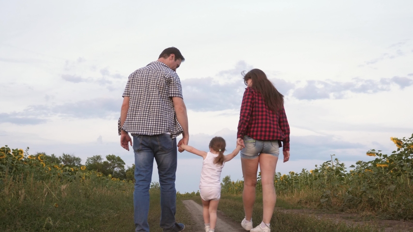 little daughter jumping holding hands mom and dad. Family with small child walks along road and laughs next to field of sunflowers.