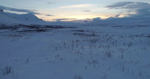 Boreal landscape, C4k aerial, tracking, drone shot close to the ground, over polar nature and snowy wilderness, arctic mountains, in the backgound, on a partly cloudy winter evening dusk, at