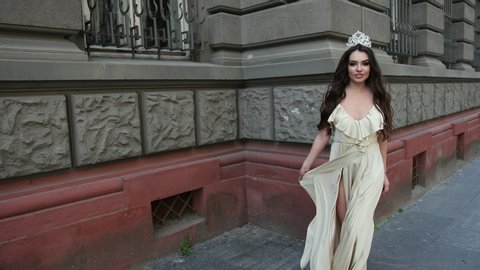 girl model, brunette, in a light long dress with a deep neckline, long hair and bright makeup, crown on his head, posing and smiling in front of the camera. photo shoot on the street near the building
