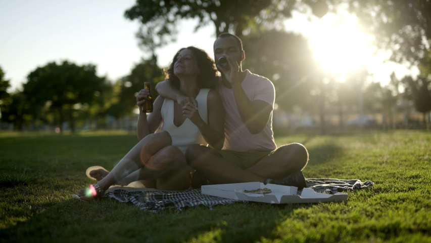 Couple with beer and pizza in park. Happy young multiethnic couple drinking beer and talking during picnic in park. Picnic concept