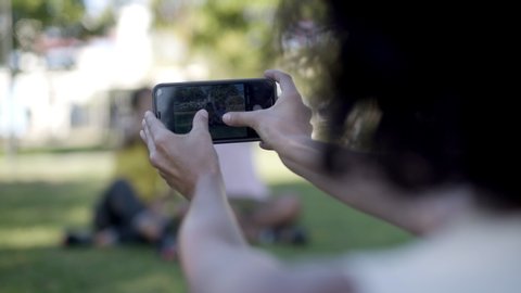 Woman photographing friends with cell phone. Cropped shot of woman holding smartphone and taking pictures of happy young friends in park, selective focus. Technology concept