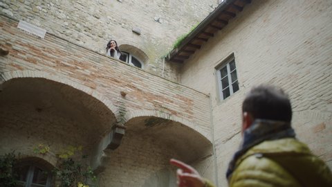Beautiful Italian woman walks out to her balcony and greets and waves to her friend as he walks into the courtyard with soft natural lighting. Medium shot on 4k RED camera.
