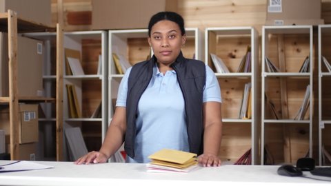 Medium portrait shot of cheerful African American woman, dressed in uniform polo shirt and bodywarmer, standing behind counter in mail service office in front of shelves and smiling for camera