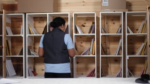 Thigh-up shot of 20-something African American female employee in uniform polo shirt and gilet picking up letters from storage shelves, returning to counter, looking towards camera and smiling