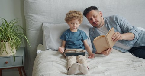 Loving father middle-aged man is reading book to cute child while kid is using tablet touching screen in bed in apartment. Family, literature and gadgets concept.