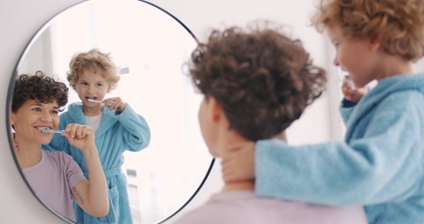 Mother and son happy family brushing teeth in bathroom looking at mirror smiling together enjoying healthcare. Boy is wearing modern bathrobe.