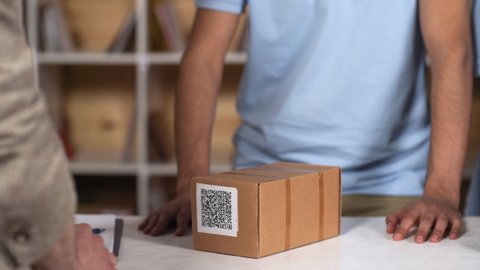 Close-up hands and body shot of dark-skinned courier service clerk in blue uniform t-shirt politely giving cardboard package with QR code to customer, who is taking it and leaving