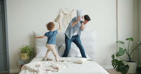 Father and little son are playing on bed fighting pillows laughing falling then mother is coming home walking in bedroom. Family, relationship and games concept.