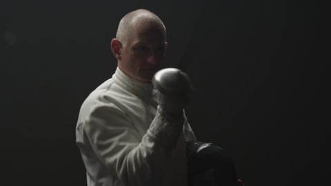 An older man fencer walks in a dark room and takes a bow with his foil