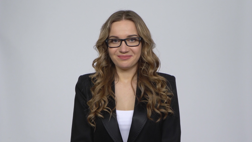 Cute girl is showing heart with fingers and blowing kiss on a gray background in the studio. The girl is wearing glasses and is wearing a black suit.