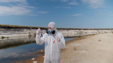 Man in a protective suit and respirator observes a chemical reaction of water in test tube, which he took for analysis from polluted river. Scientist takes samples in vitro. Ecological disaster. 4K