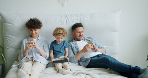 Slow motion of mother and father young people using modern smartphones while child small boy is holding tablet in bed. Gadget addiction and technology concept.