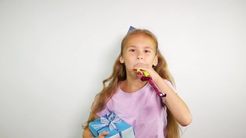 Birthday kid holds a gift and blows noisemaker. Lovely young girl wearing birthday cap over white background