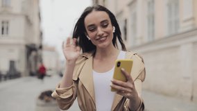 Beautiful Happy Young Girl Wearing Wireless Earphones Making a Video call Smiling Talking Waving her Hand Walking Relaxed on the Street City Background Close Up.