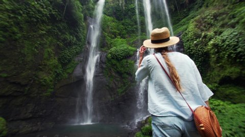 Back of woman woman in straw hat enjoying breathtaking high waterfall Sekumpul in Bali rainforest. Great wild nature of Indonesia