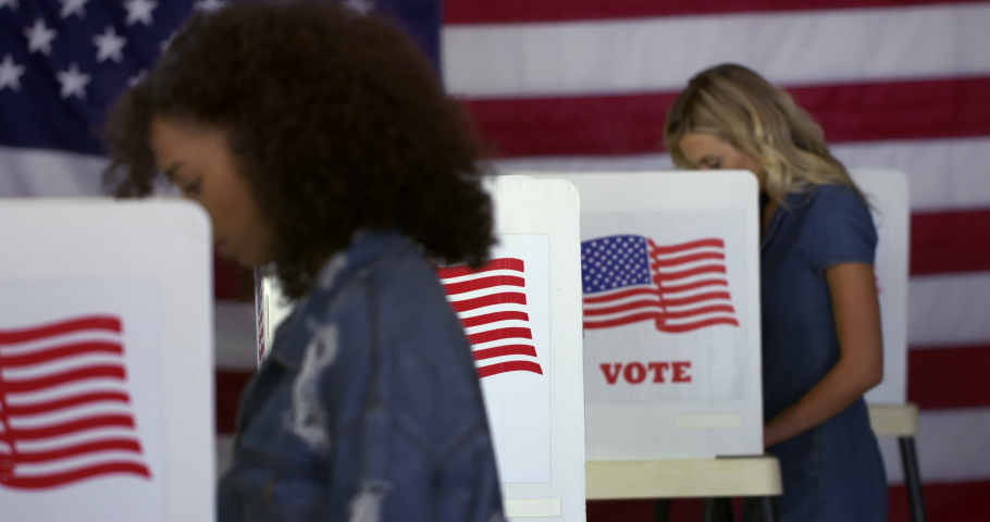 MS Hispanic man enters polling station and votes in a booth with young Hispanic woman in foreground and blonde Caucasian woman behind and US flag in background. Real time 4K Royalty-Free Stock Footage #1035292043