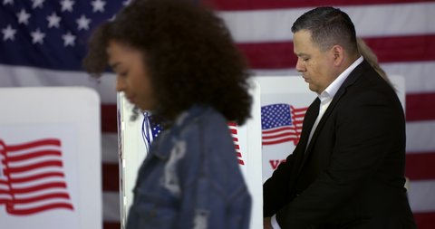 MS Hispanic man enters polling station and votes in a booth with young Hispanic woman in foreground and blonde Caucasian woman behind and US flag in background. Real time 4K