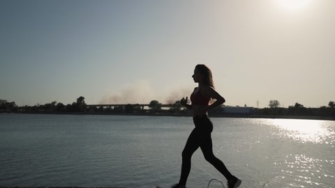 Young athletic caucasian girl runs along the lake on a sunset background. Far away smoke from the fire. Slow motion. Overall plan.