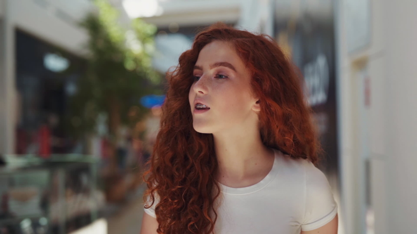 Gorgeous curly redheaded girl excitedly looks around while walking around the big mall with many stores. Consumerism and lifestyle concept. Positive emotions, good mood, facial expression. Female