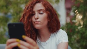 Close up view of extremely beautiful redheaded curly student girl smiling cutely, stroking her gorgeous hair while taking selfie on green plants background. Social networks addiction, technologies