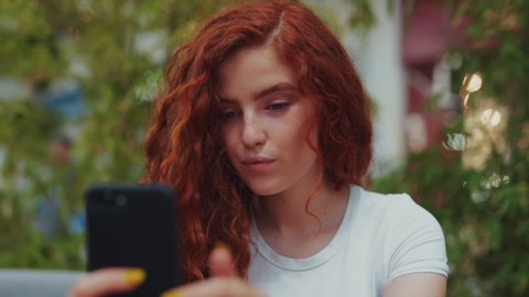 Close up view of lovely curly ginger student girl sitting in café alone and actively chatting with her friends, followers via smartphone. Online, browsing social networks, communication and