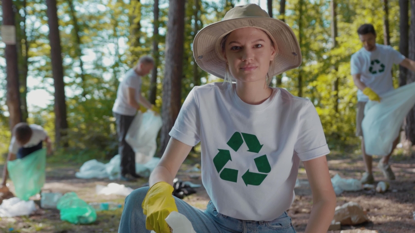 Close up young woman with hat cleans forest of debris folding it in plastic bag look at camera smile  | Shutterstock HD Video #1035318224