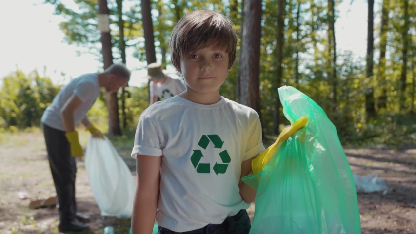 Cute boy voluteers activists child in gloves tidying up rubbish in park or forest look at camera smile | Shutterstock HD Video #1035318251