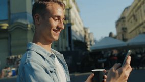 smiling young man walks along city street holding coffee cup and swiping photos on telephone close view slow motion - Powered by Shutterstock - Get 15% off with code: PIKWIZARD15