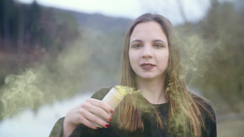 Girl wave with yellow smoke. Static slow-motion shot of a female person in focus looking into the camera and holding a smoke bomb in one hand. Background lake and trees.