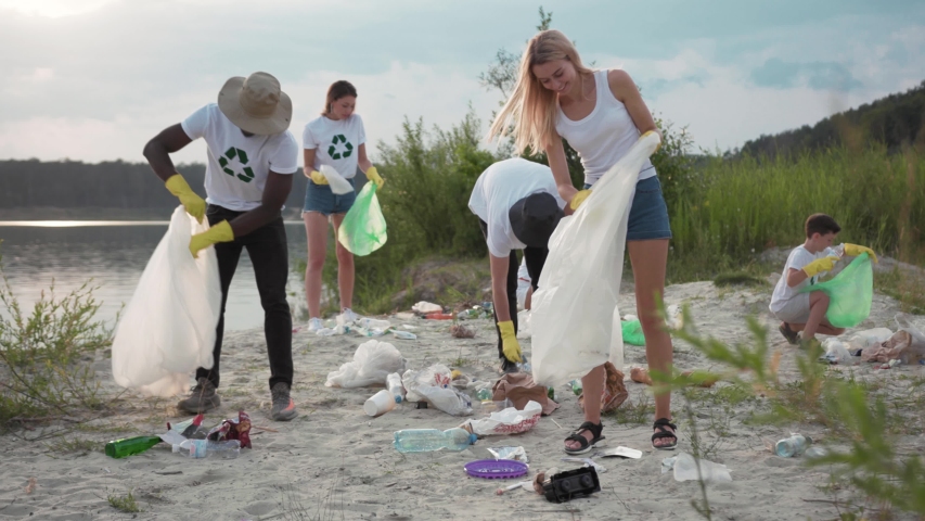 Volunteer young team cleaning up the dirty beach from plastic carrying bags to collect trash on sunny day at a lake. Children and adults caring about the environment. Royalty-Free Stock Footage #1035365405