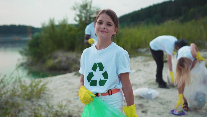 Porrtrait of little cute cheerful girl cleaning the sandy beach from garbage smiling of joy having good time outdoor. Young volunteer team on eco-friendly movement. | Shutterstock HD Video #1035365459