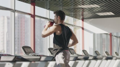 Runner man drinking water from bottle running on treadmill machine. Athlete man drinking water practicing on running machine. Handsome guy opening bottle with water in sport club.