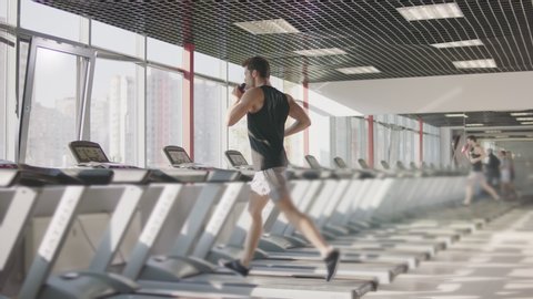 Running man drinking water on treadmill machine in gym club. Thirsty man sipping liquid at cardio training on running machine. Back view of handsome runner training with bottle on treadmill.