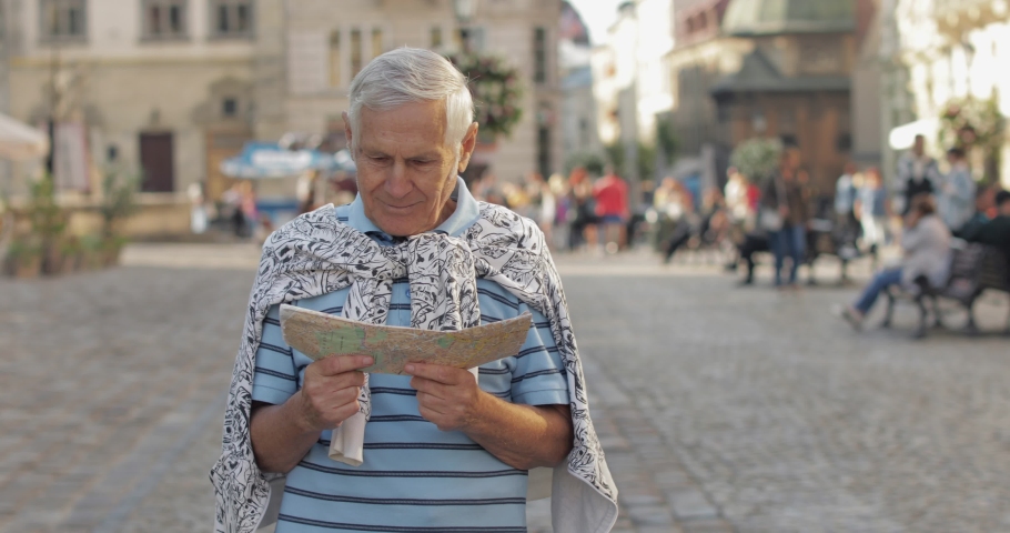 Senior male tourist exploring town center with a map in hands and looking for the route while traveling in Lviv, Ukraine. Smiling. T-shirt and sweater in his neck. Vacation concept