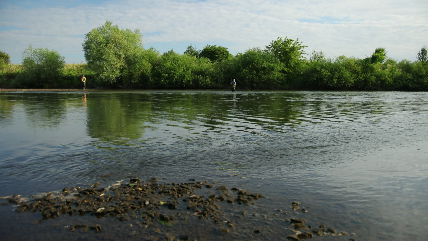 Angling, two men in special clothes holding fishing rods, two people fishing on the river, standing in the water, a small current, the nature is beautiful, summer day, Wide angle, slow motion
