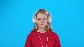 Girl listening to music in big white headphones, singing and dancing on blue background. Teen enjoying music. Slow motion