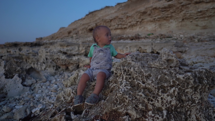 Kid sitting on the stones on the sea coast at sunset. Boy talking to himself and fidgeting