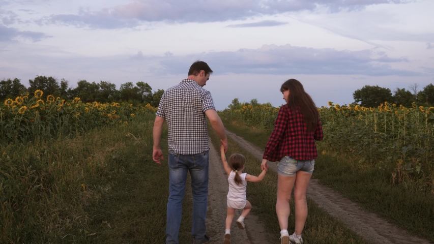 little daughter jumping holding hands mom and dad. Family with small child walks along road and laughs next to field of sunflowers. Mom, dad and daughter are resting together outside city in nature.
