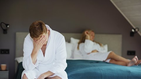 Frustrated middle aged man in white bathrobe sitting on bed after sexual failure or adultery and rubbing forehead, female partner lying in background. Man raising head and looking at camera