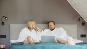 Married middle aged couple in white bathrobes lying on bed in hotel room, relaxing and talking. Man using laptop computer - Powered by Shutterstock - Get 15% off with code: PIKWIZARD15