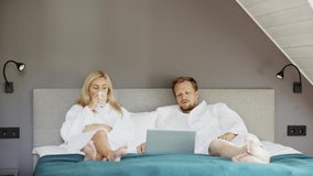 Married middle aged couple in white bathrobes lying on bed in hotel room, relaxing and talking in morning. Man using laptop computer, woman drinking hot tea or coffee - Powered by Shutterstock - Get 15% off with code: PIKWIZARD15