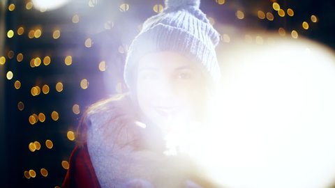 Girl play with sparkler surrounded by Christmas light. Portrait sliding shot of young woman in focus wearing winter clothes and surrounded by decorative lights.