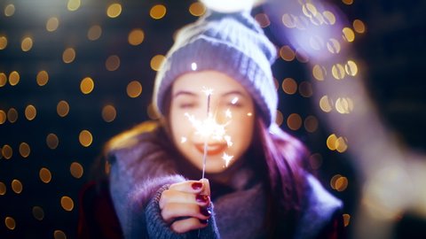 Girl holding sparkler enjoying Christmas spirit. Portrait sliding shot of young woman in focus wearing winter clothes and surrounded by decorative lights.